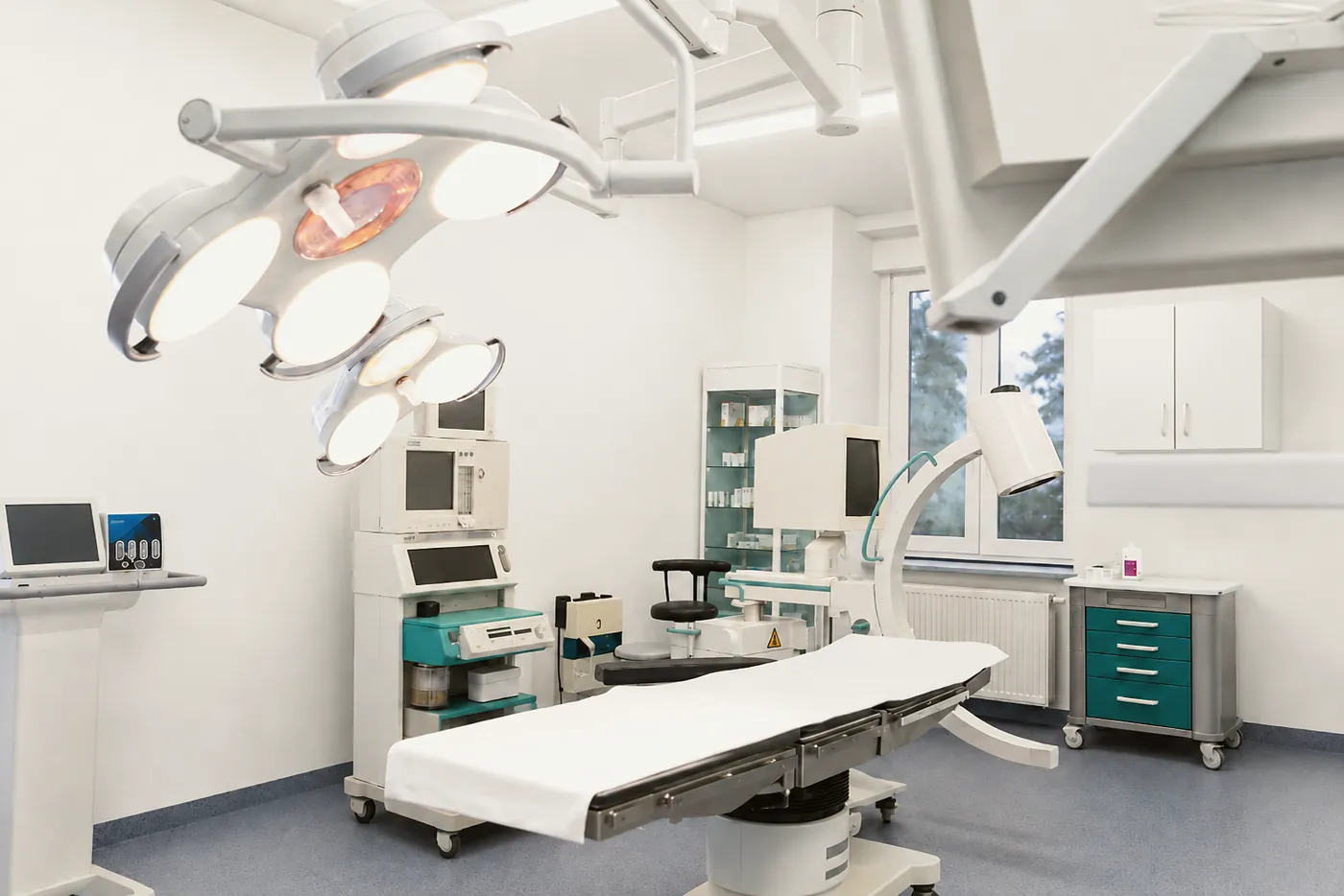 Stół operacyjny oświetlony lampami bezcieniowymi, obok aparatura monitorująca i szafa z materiałami.